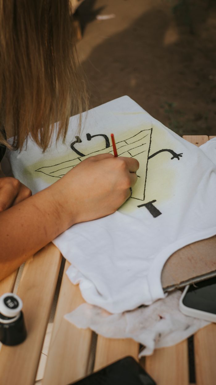 A woman creatively paints a unique design on a white t-shirt outdoors, enhancing the garment's style.