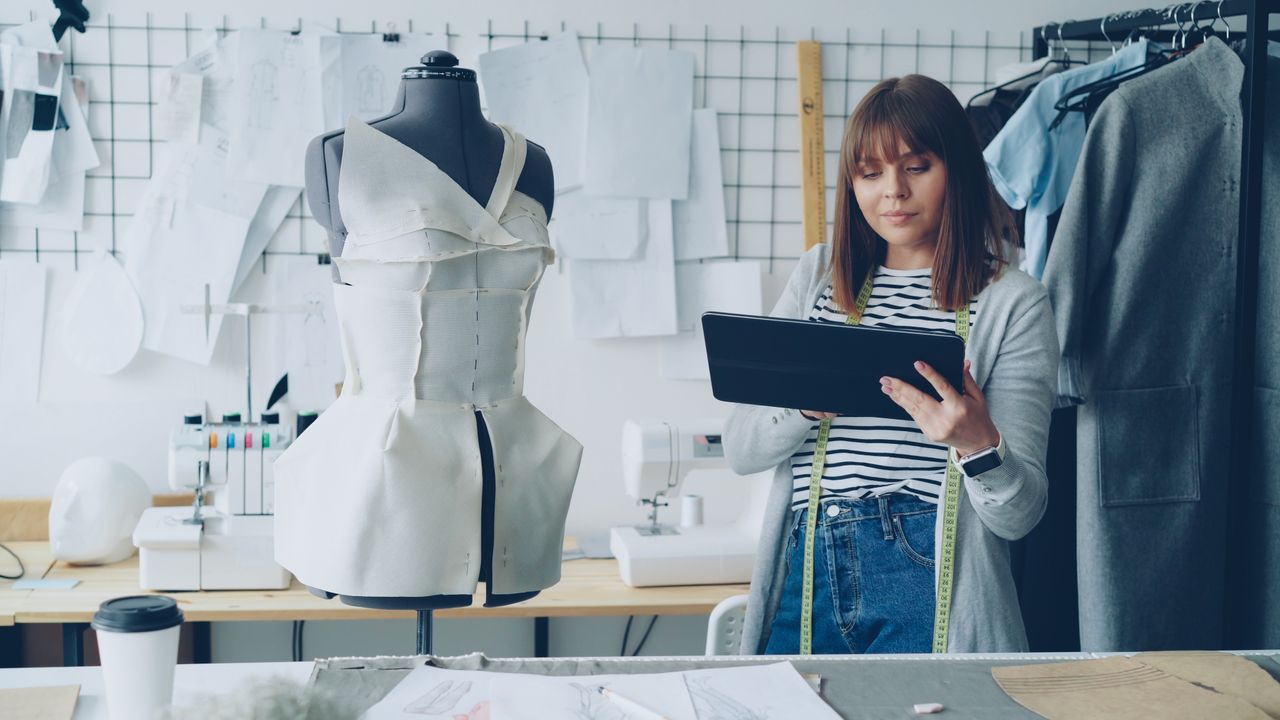 A fashion designer works with a tablet and dress form in a creative studio space.