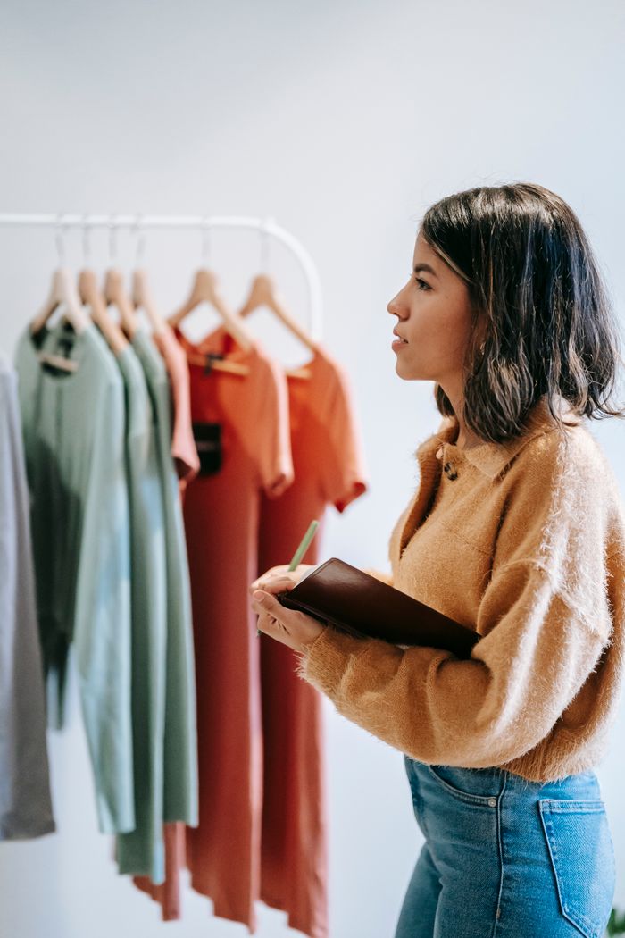 Home Side view of female seller in casual outfit standing near wardrobe with various multicolored clothes on hangers with notebook in light store